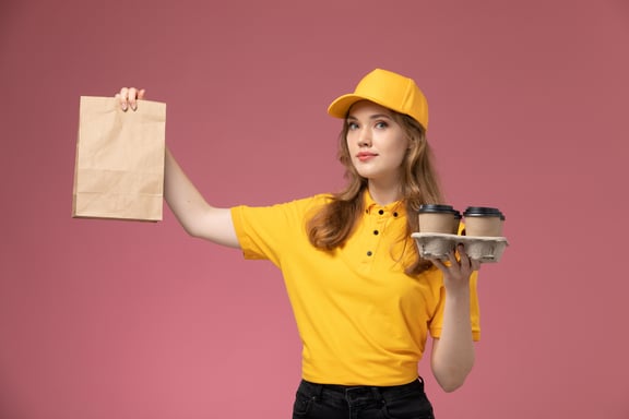 front-view-young-female-courier-yellow-uniform-holding-coffee-cups-package-with-food-pink-desk-desk-job-uniform-delivery-service-worker