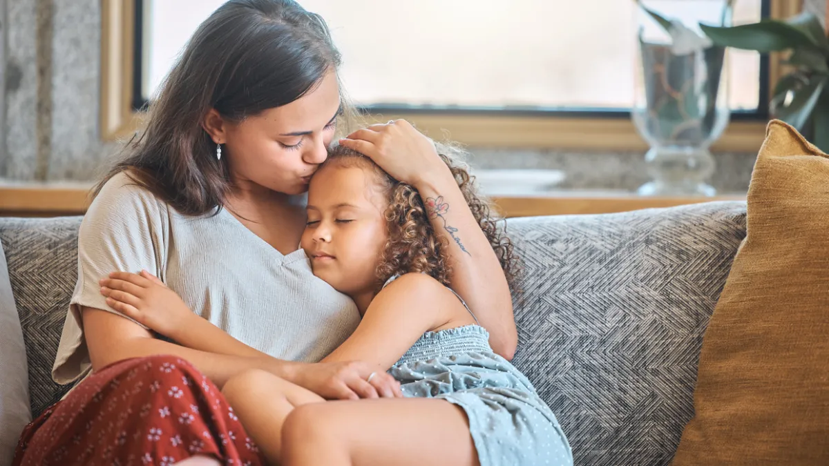 A woman holds a young child close and kisses her forehead as they sit together on a couch.